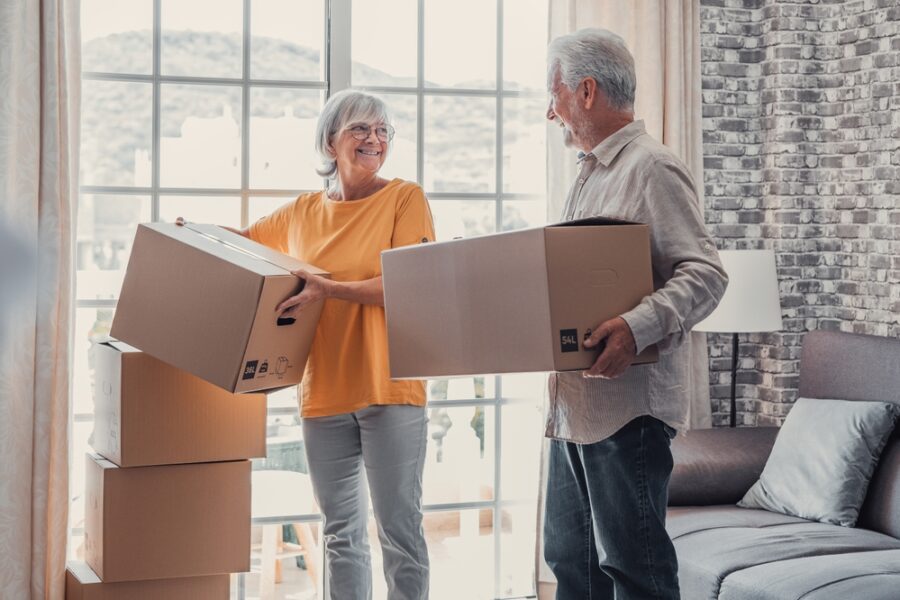 Two older adults, each carrying a box, as they smile to each other as they prepare to move