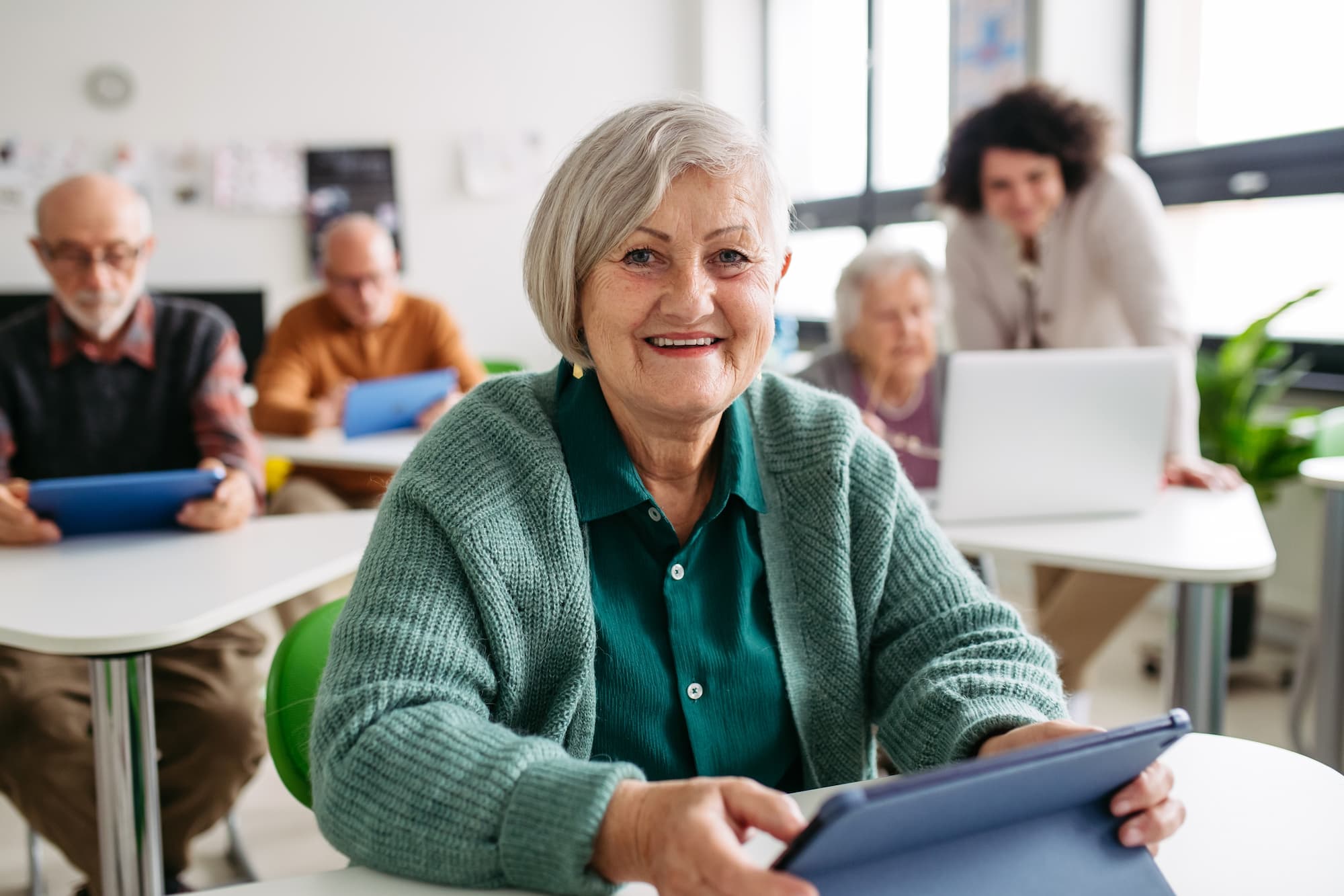 A senior woman sitting in a classroom desk with her blue ipad learning about online safety, with other older adults in the background learning