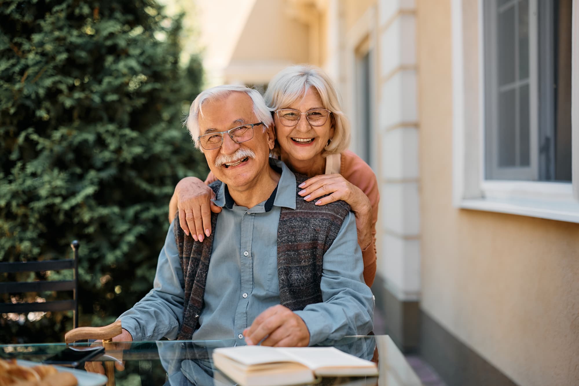 An elderly man and elderly woman who's resting her hands on his shoulder, smiling together towards the camera